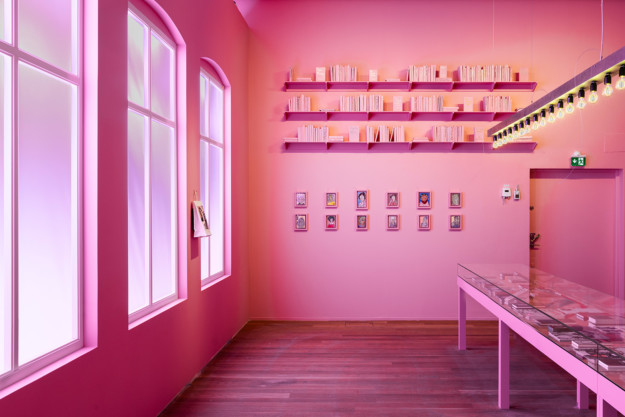 Installationsvy i rummet "The Wicked Pavilion". Ett rum helt i rosa, med en monter och hyllor med böcker på väggen i bild. Under hyllorna hänger målningar, och brevid väggen syns tre fönster i kyrkostil. 
