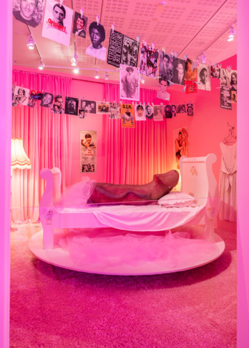 Installationsvy i rummet "The Wicked Pavilion". Fotografi av ett flickrum i rosa. I taket hänger en fisklina med fotografier. I mitten av rummet står en säng på ett podie, i sängen ligger en stor fallos-skuptur. 