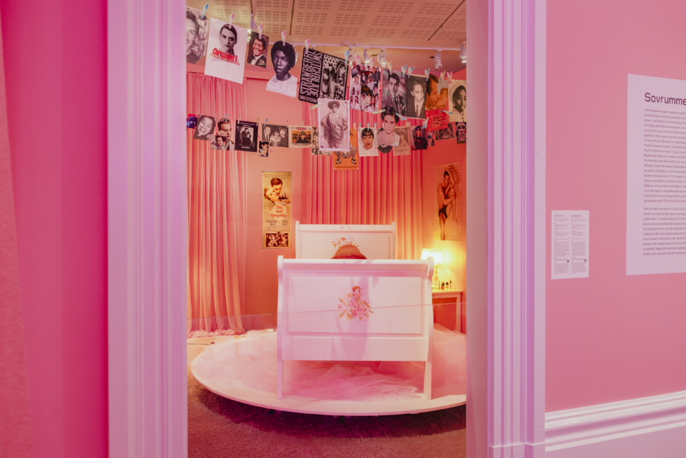 Installation view from the room "The Wicked Pavilion". Photograph of a girl's room in pink. A fishing line with photographs hangs from the ceiling. In the middle of the room is a bed on a podium, in the bed is a large phallus sculpture.